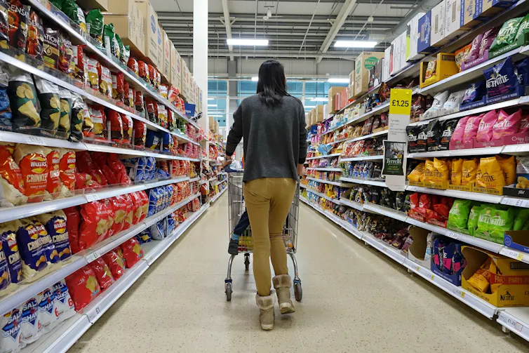 woman pushes a shopping trolley with a child in the seat down a supermarket aisle.