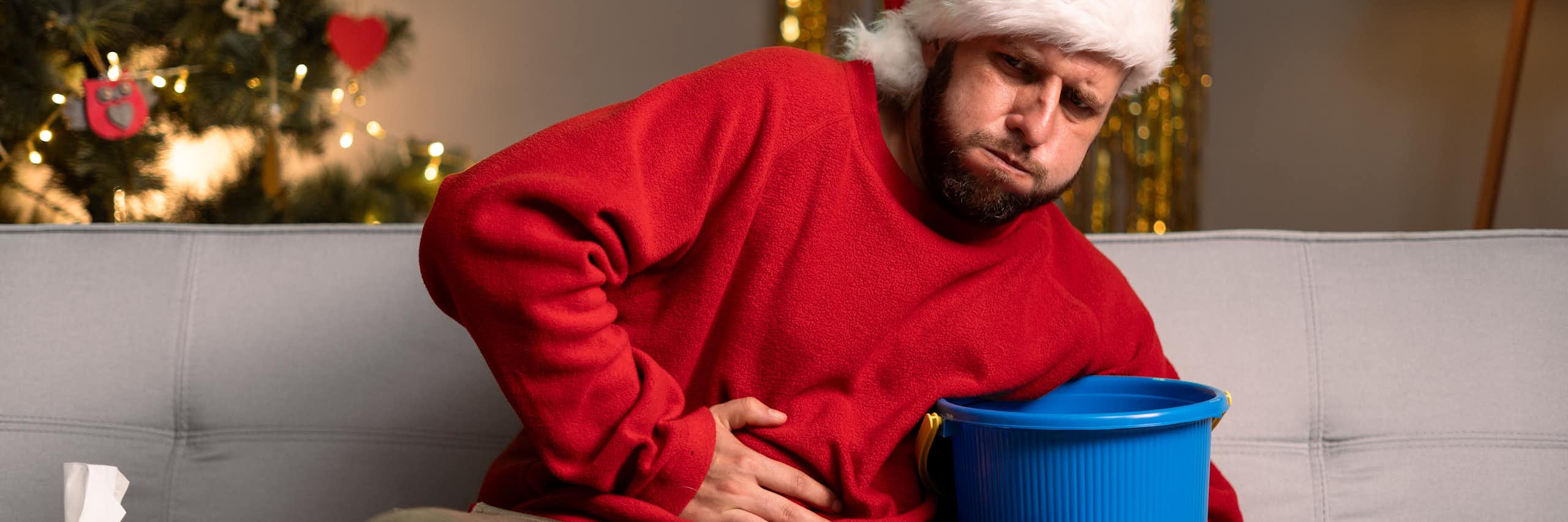 Homem sentado no sofá, vestindo um suéter natalino e um chapéu de Papai Noel, com uma árvore de Natal ao fundo, segura um balde para vômito com uma mão e segura o estômago com a outra.