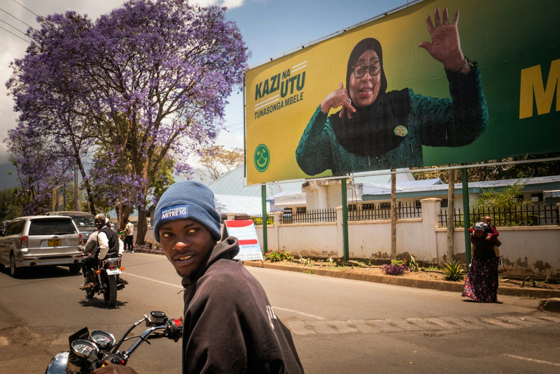 A man on a motorcycle looks over his shoulder as a large political billboard can be seen behind him.