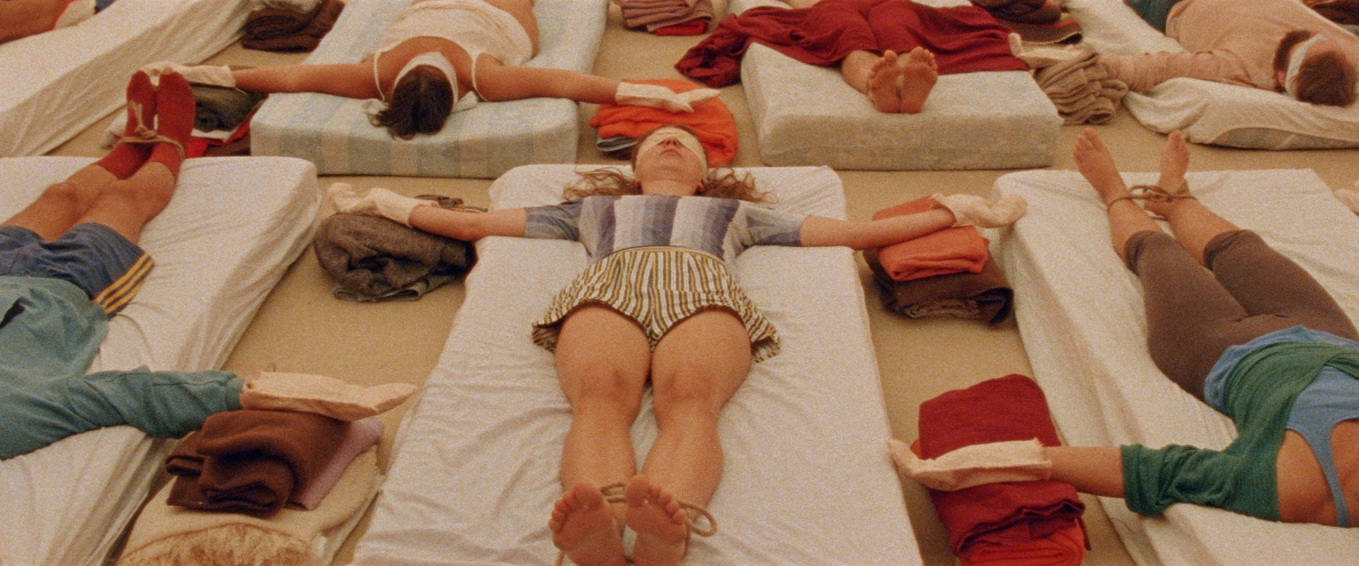 Plano cenital de varias personas tumbadas en colchones en el suelo, con los ojos vendados y las manos y pies atados, participando en una terapia grupal.