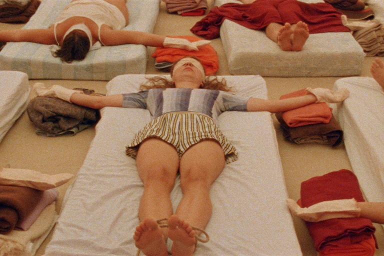 Plano cenital de varias personas tumbadas en colchones en el suelo, con los ojos vendados y las manos y pies atados, participando en una terapia grupal.