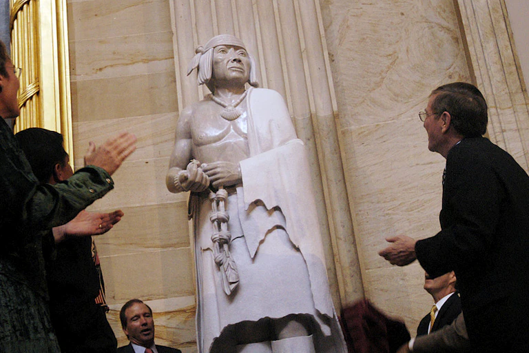 A stone statue of a man with a necklace and head covering, with a blanket thrown over his shoulder, stands in a tall room as men around it clap.