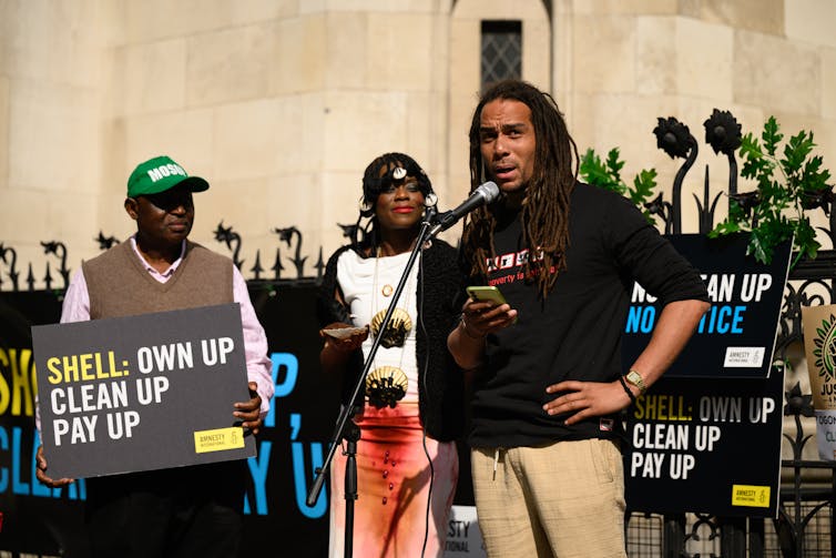 A man speaks into a microphone. Behind him, a person holds a sign reading: 'Shell: Own up, clean up, pay up'
