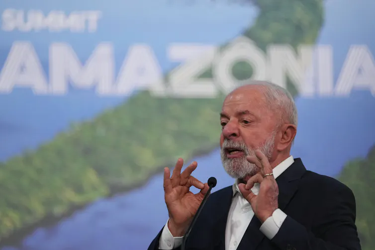 O presidente Lula gesticula com as mãos enquanto fala em frente a uma foto da Amazônia.