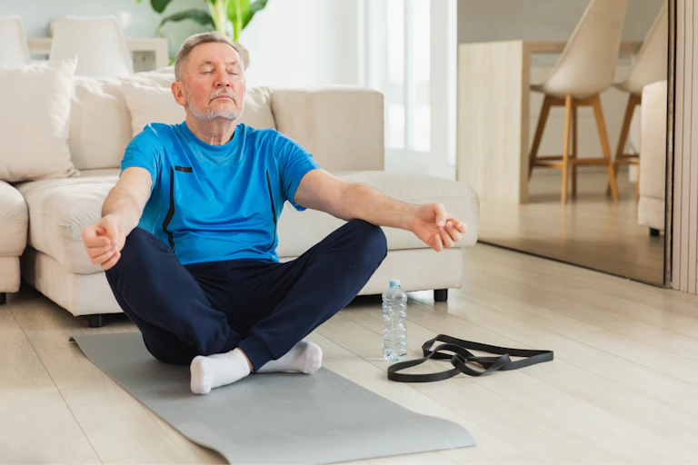 Un monsieur d'un certain âge est assis en position de yoga dans son salon.