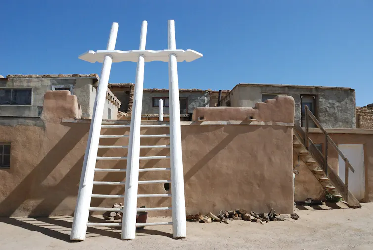 A large wooden ladder with three poles, painted white, leans against an adobe wall under a bright blue sky.