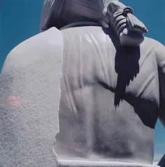 A stone statue of a man with a ponytail and a blanket thrown over one shoulder, seen from the back.