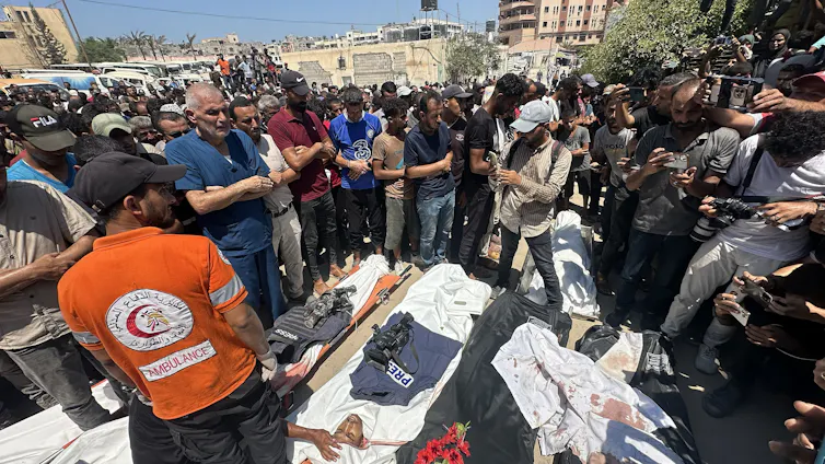 A crowd of people around three bodies covered in white sheets and press flak jackets on the ground