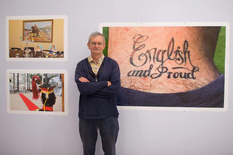 A grey-haired casually dressed man standing in front of his photographs in a gallery exhibititon.