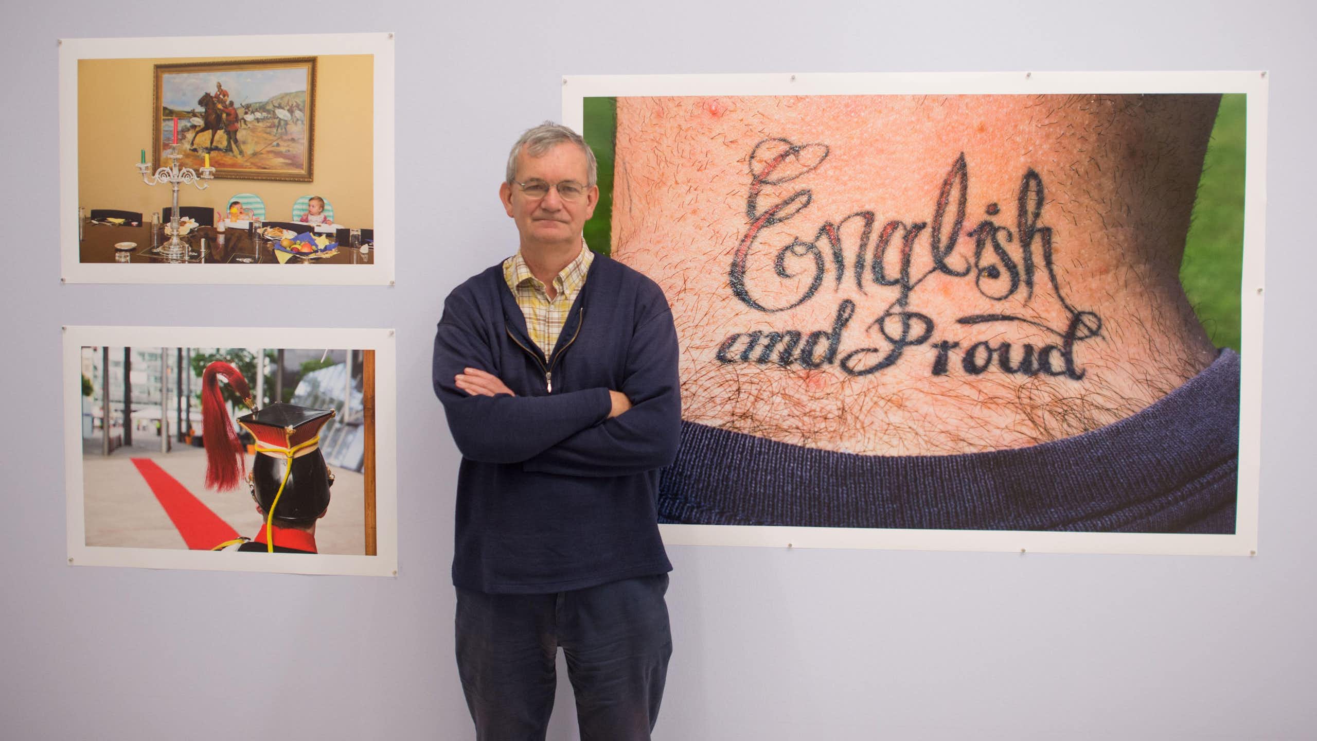 A grey-haired casually dressed man standing in front of his photographs in a gallery exhibititon.