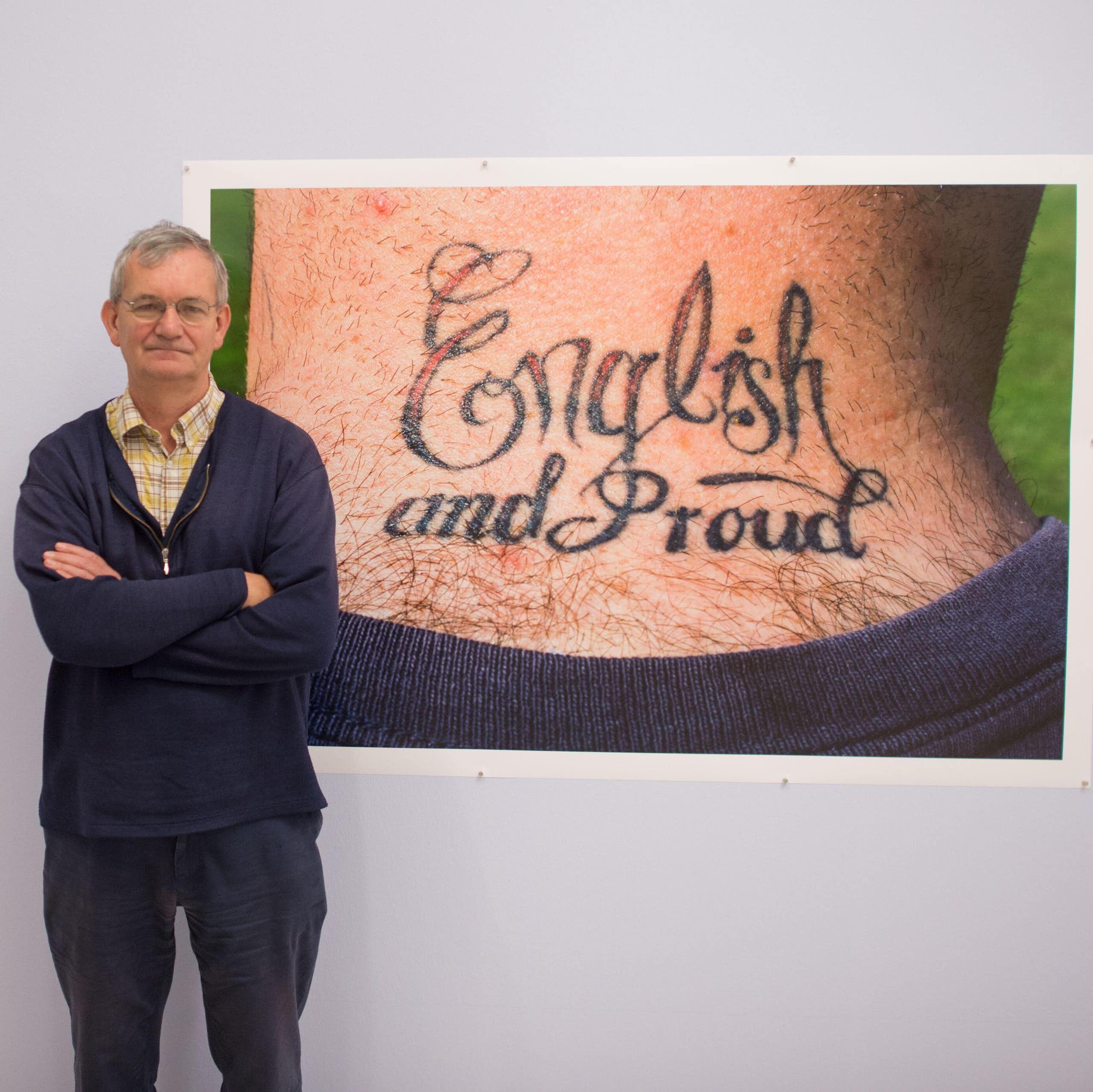 A grey-haired casually dressed man standing in front of his photographs in a gallery exhibititon.