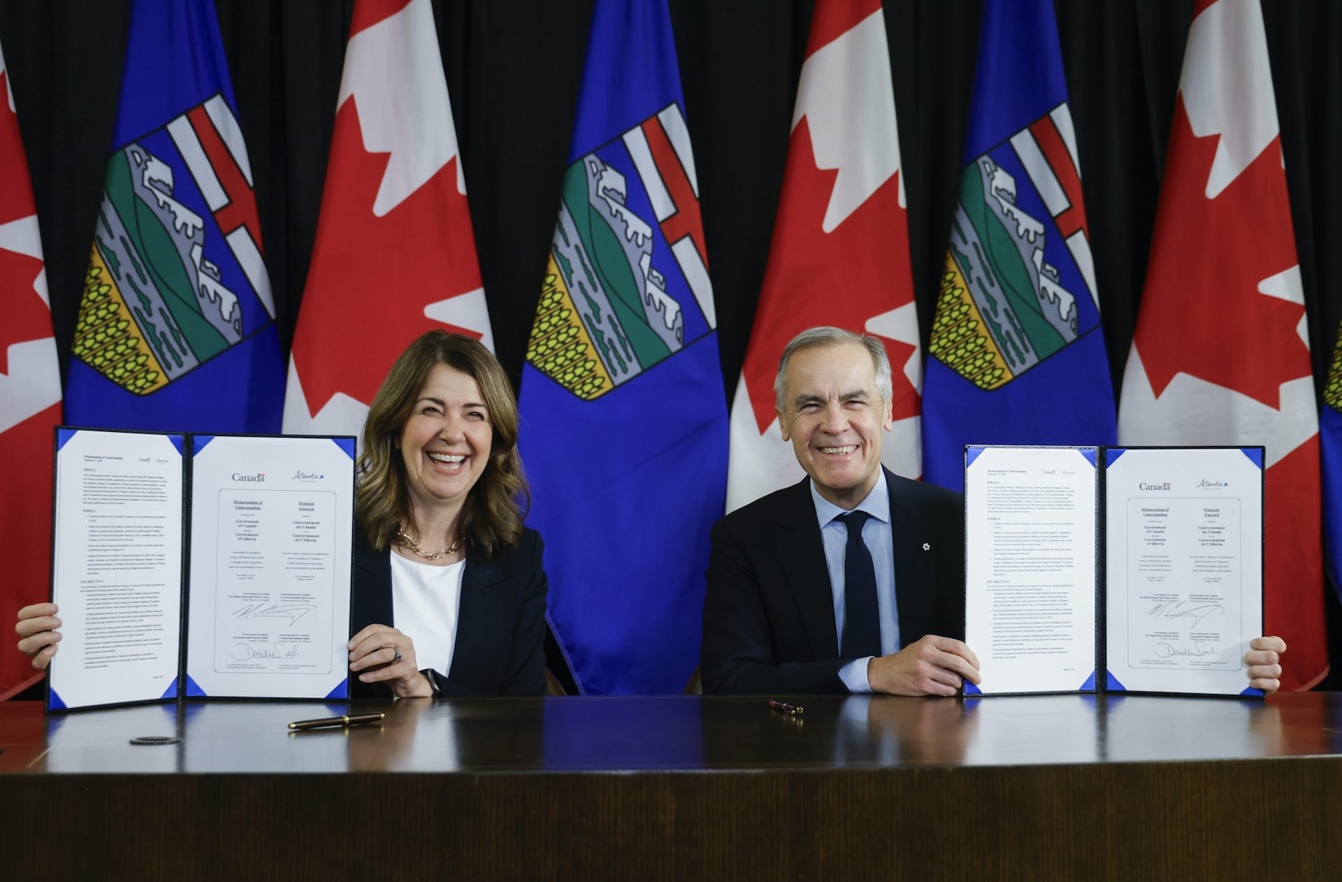 Un hombre y una mujer vestidos con ropa de negocios están sentados en una mesa con documentos. Detrás de ellos están las banderas de Canadá y Alberta en postes.