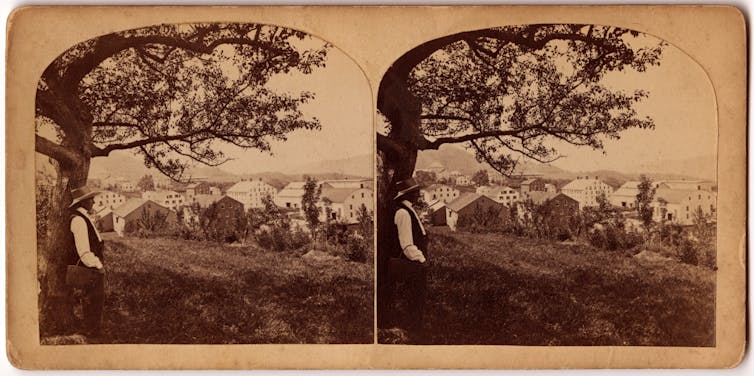 Imagen en tonos sepia de un hombre con camisa blanca, chaleco oscuro y sombrero de paja parado bajo un árbol en la cima de una colina que domina muchas casas.