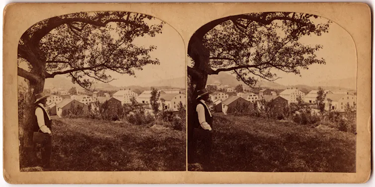 A sepia-toned image of a man in a white shirt, dark vest and straw hat standing under a hilltop tree overlooking many houses.