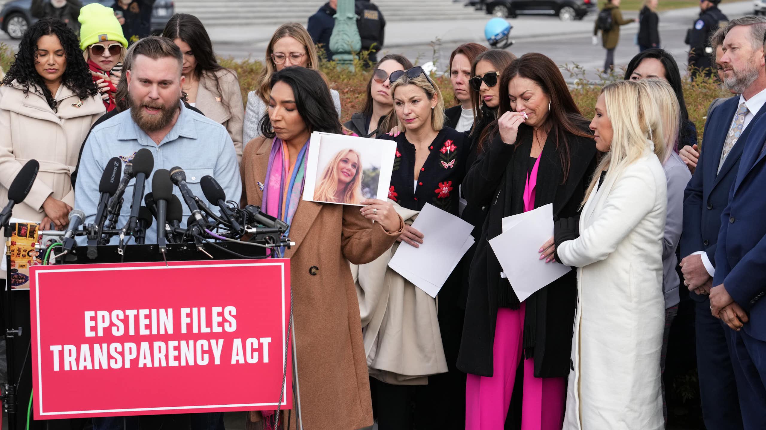 a man stands before a bunch of mics and speaks as a crowd of women, some holding pictures, stand behind him, some in tears