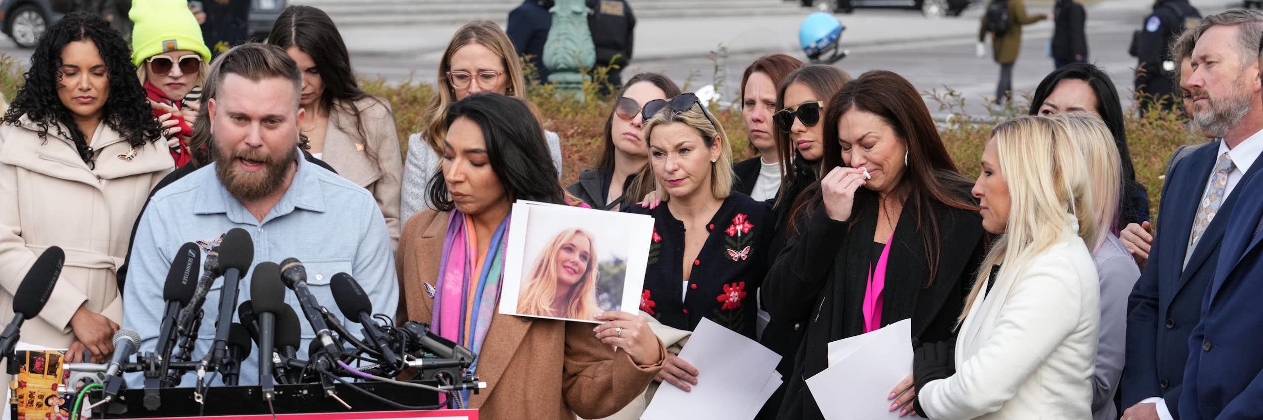 a man stands before a bunch of mics and speaks as a crowd of women, some holding pictures, stand behind him, some in tears