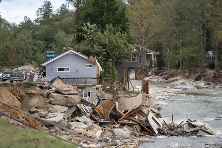 Two homes at the river's edge, one missing half of it's lower floor, with top hanging over precariously. Lots of boulders and debris are in the water.
