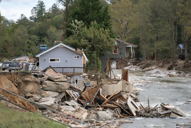 Two homes at the river's edge, one missing half of it's lower floor, with top hanging over precariously. Lots of boulders and debris are in the water.