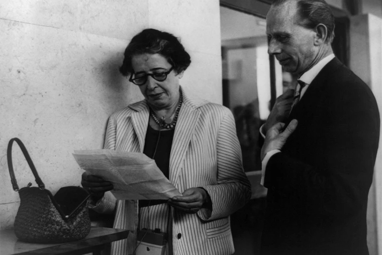 A woman in a white suit and glasses reading some papers next to a newpaper reporter.