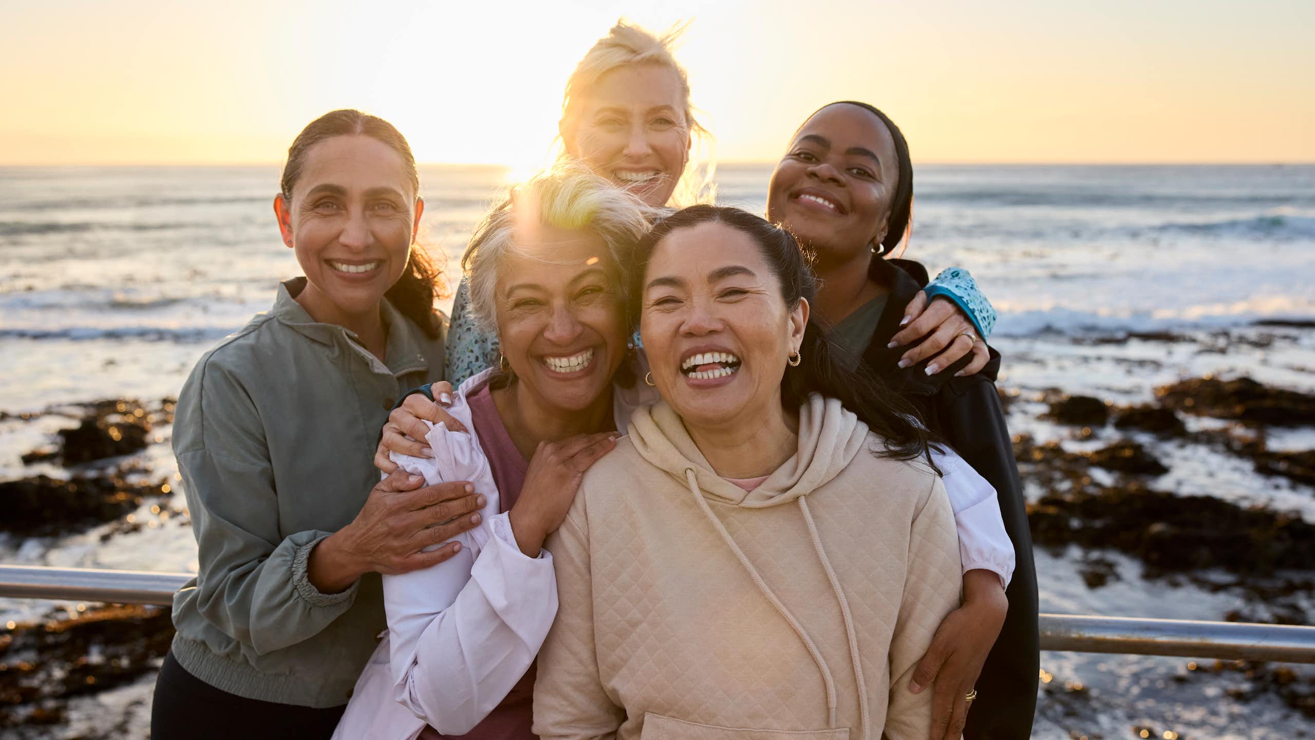 Five female friends posing with each other on a beach against sunny sky.