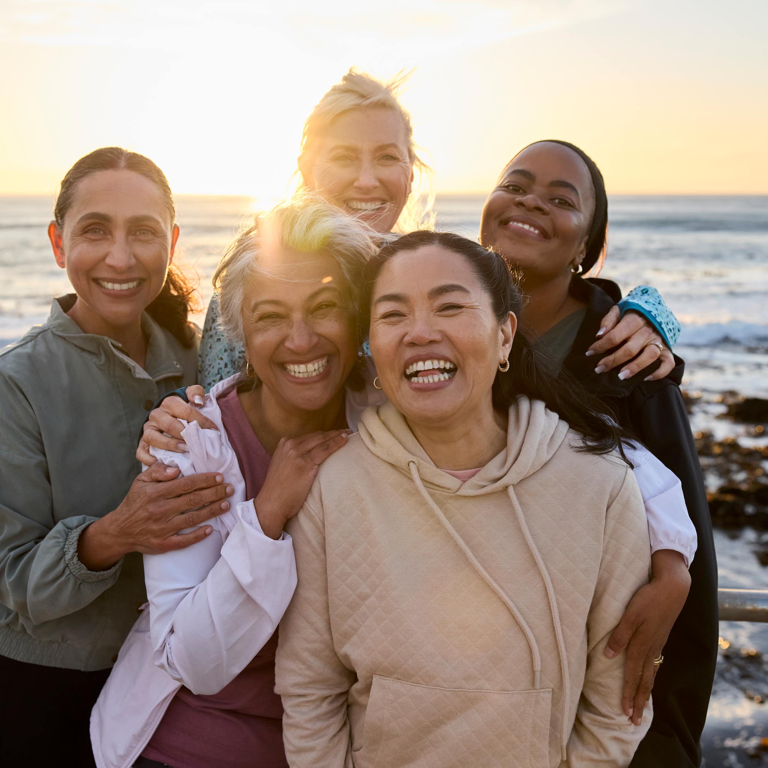 Five female friends posing with each other on a beach against sunny sky.