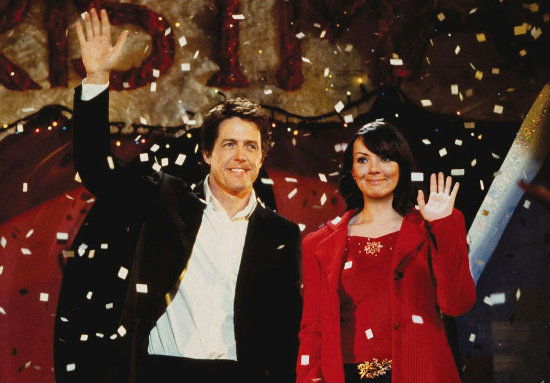 Un hombre y una mujer saludan y sonríen desde un escenario.