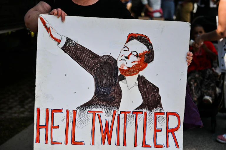 A protester holds up a sign featuring an illustration of Elon Musk and the words 'heil twittler'.