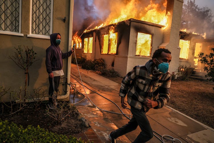 A man turns a hose on a burning house while another runs.