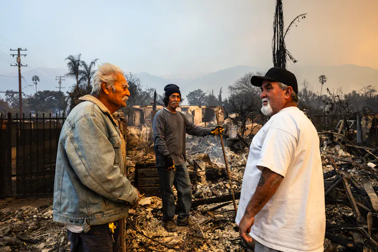 Three men, one an older man, stand in the still-smoky ruins of what was once a home, with fire damage all around them.