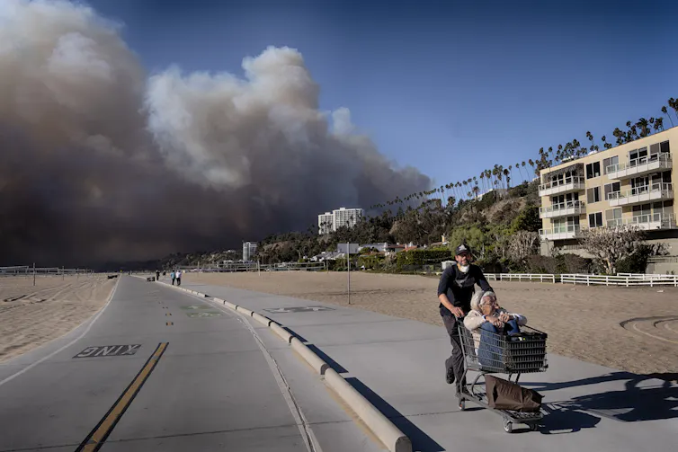 A man pushes an older woman in a shopping cart along a pathway with apartments on one side and sand on the other, and thick smoke behind them.