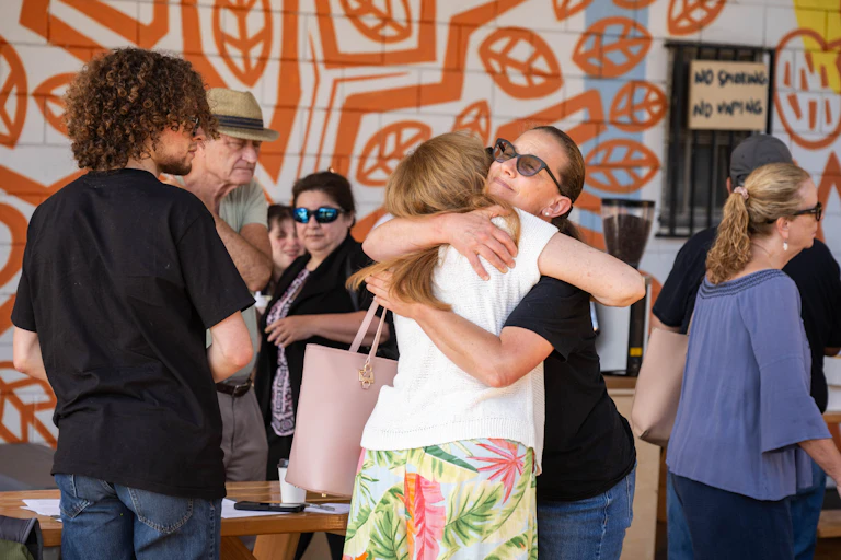 Two women hug with a group of neighbors from all ages around them.