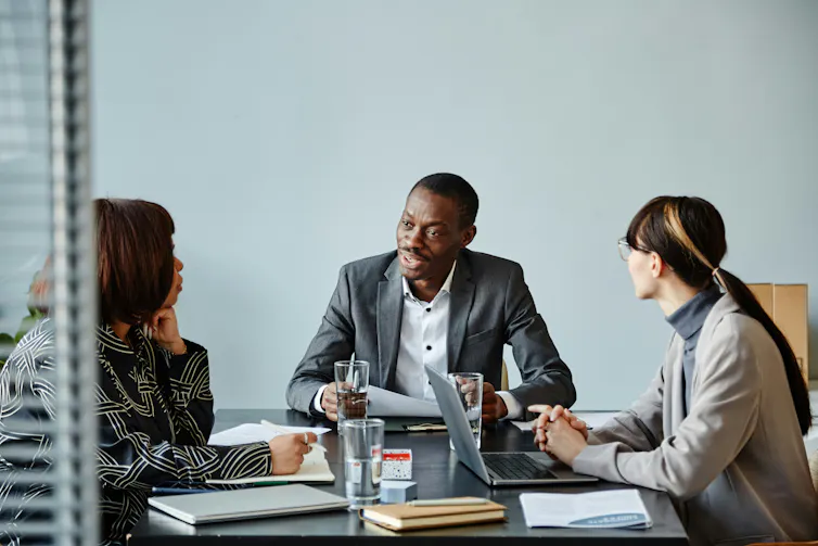 Two women speak to a man at a conference table in an office
