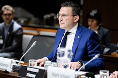 Un hombre de pelo oscuro y traje azul está sentado detrás de un micrófono.