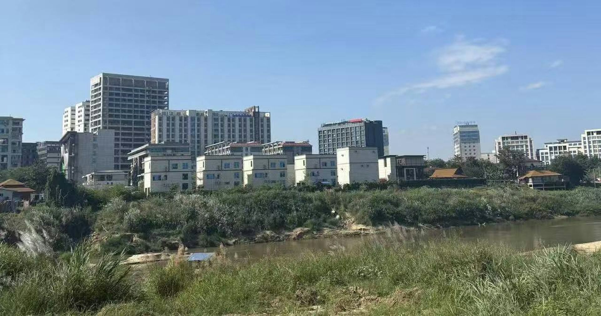 View across the Moei River towards Shwe Kokko with the river and grassy riverbank in the foreground under a clear blue sky.