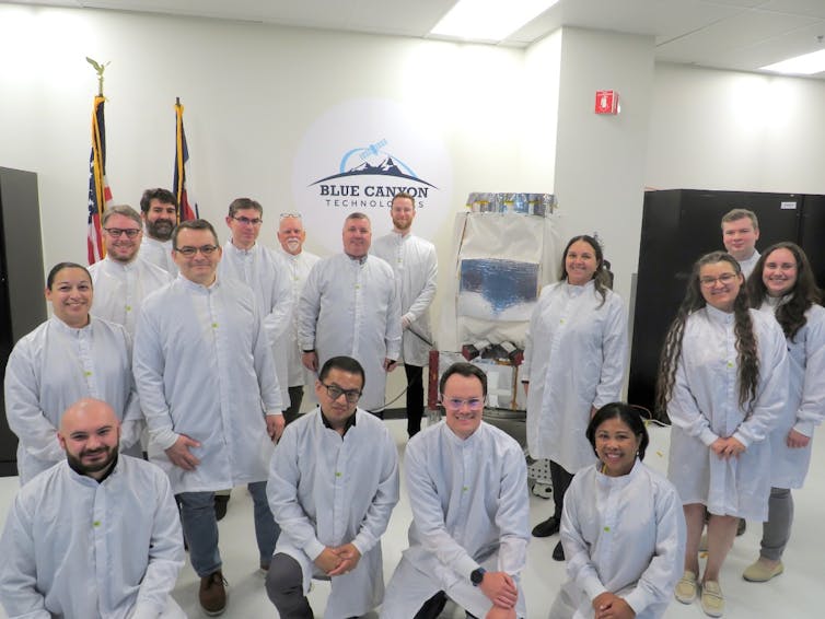 Un grupo de personas con batas de laboratorio, de pie junto a un satélite.