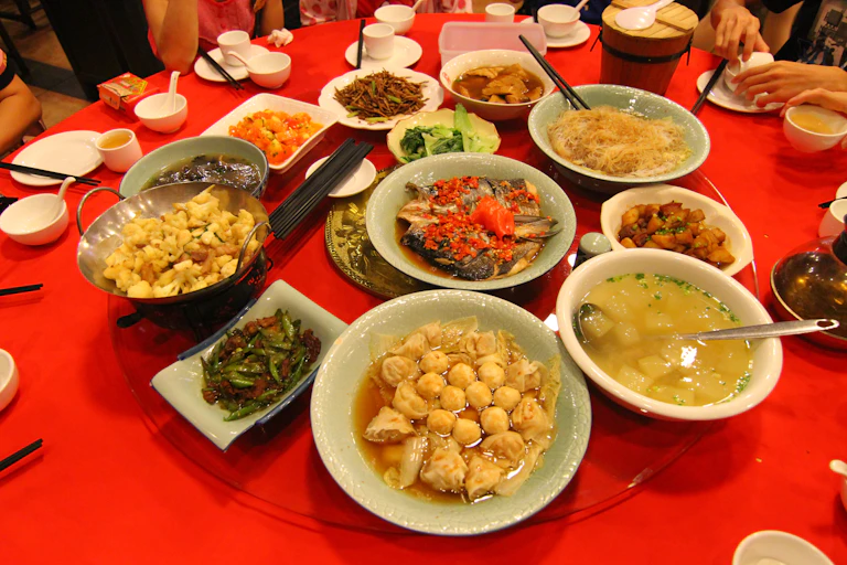 Plates of chinese food including dumplings, green beans, soup and sauces in bowls set on a red tablecloth