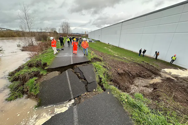 A paved bicycling path atop a levee is broken and slabs of asphalt pavement are tilted into a breach where water poured through.