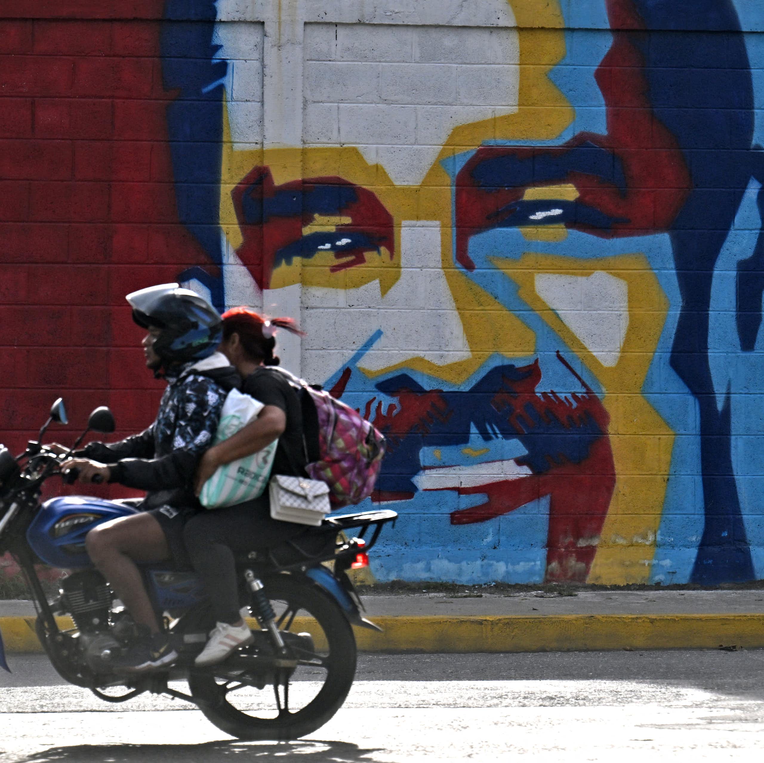 A man on a motorcycle drives past a mural of a man's face.