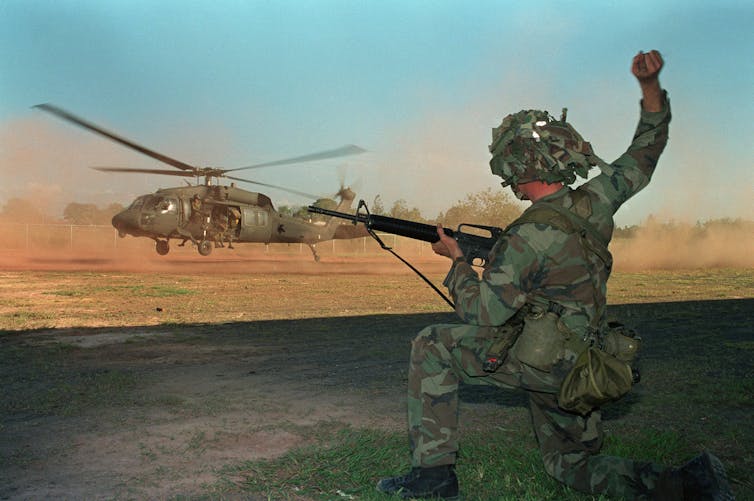 A man in army fatigues is in front of a landing helicopter