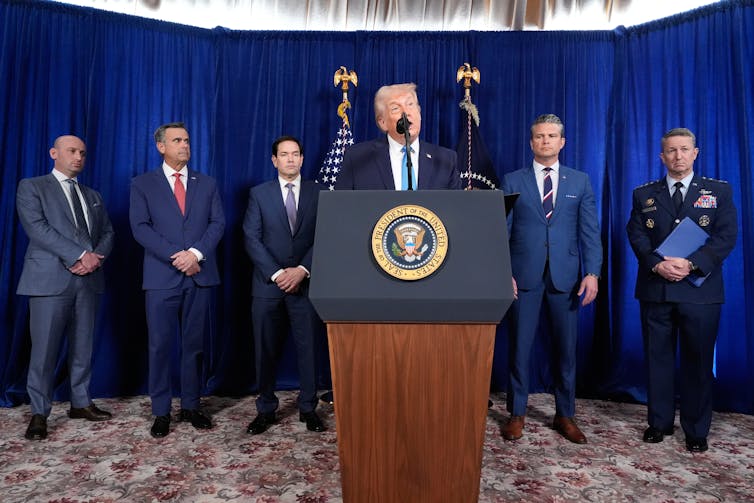 a white man in a suit stands at a podium with the presidential seal, while several other men stand behind him