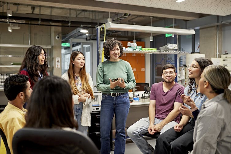 A group of young people stand in a circle in what looks like a science lab.