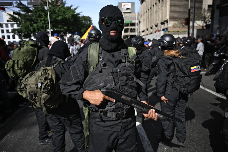 A man in a mask carrying a gun is flanked by others dressed in black