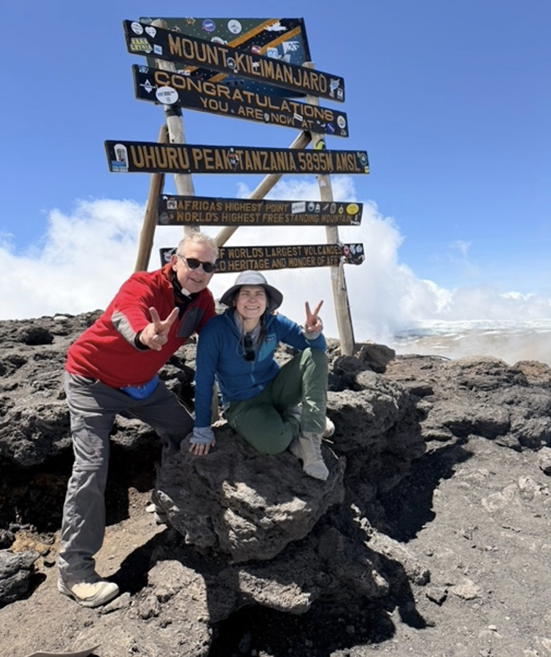 Dos personas riendo bajo un cartel en el monte Kilamanjaro. Felicitaciones, ahora estás en la cima de Uhuru, Tanzania.