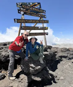 Two people smiling at beneath a sign reading Mount Kilamunjaro Congratulations you are now at Uhuru Peak, Tanzania