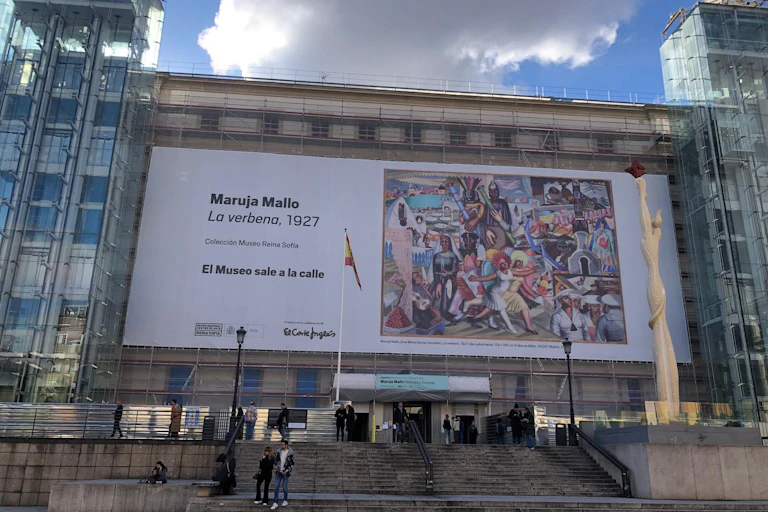 Fachada de un museo con una lona que muestra un cuadro colorista de Maruja Mallo que retrata una verbena.
