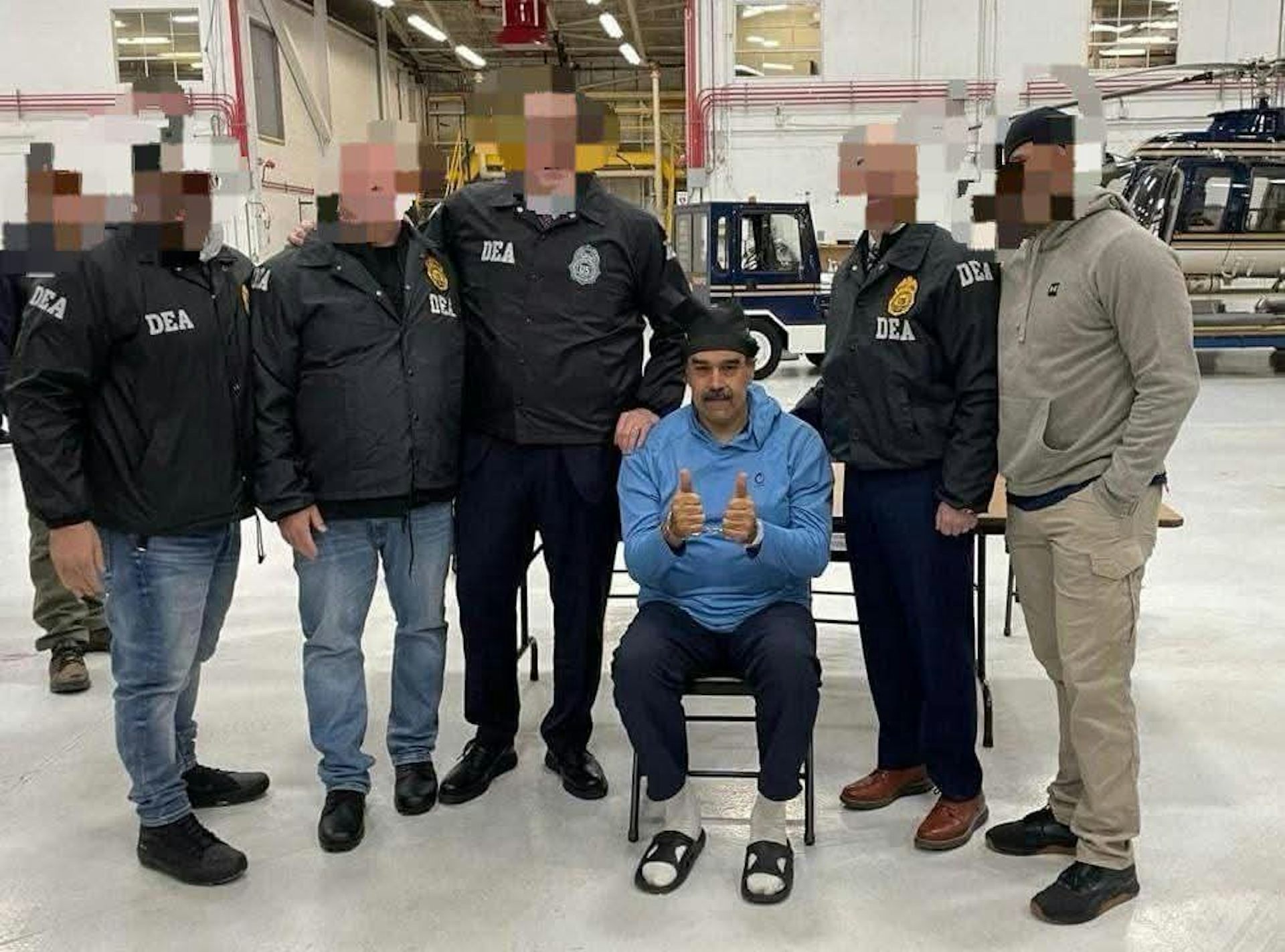 Nicolás Maduro, handcuffed and wearing prison clothes, with Drug Enforcement Agency officers with theiur faces obscured.