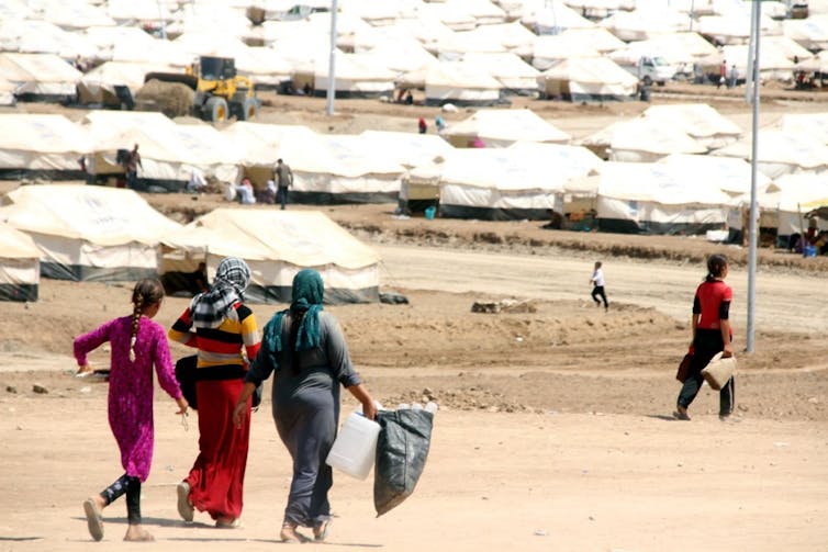 Three women walk towards lines of tents in a refugee camp.