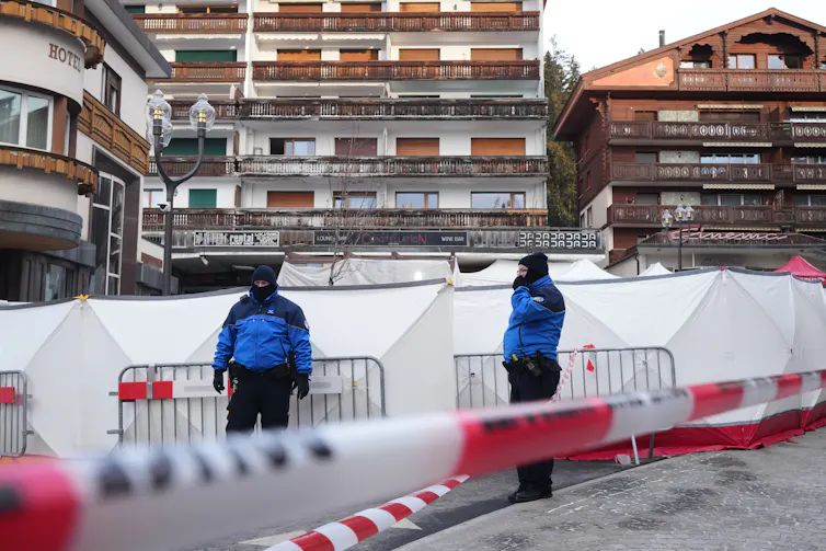 two men in winter jackets stand in front of white tents and metal barricades.