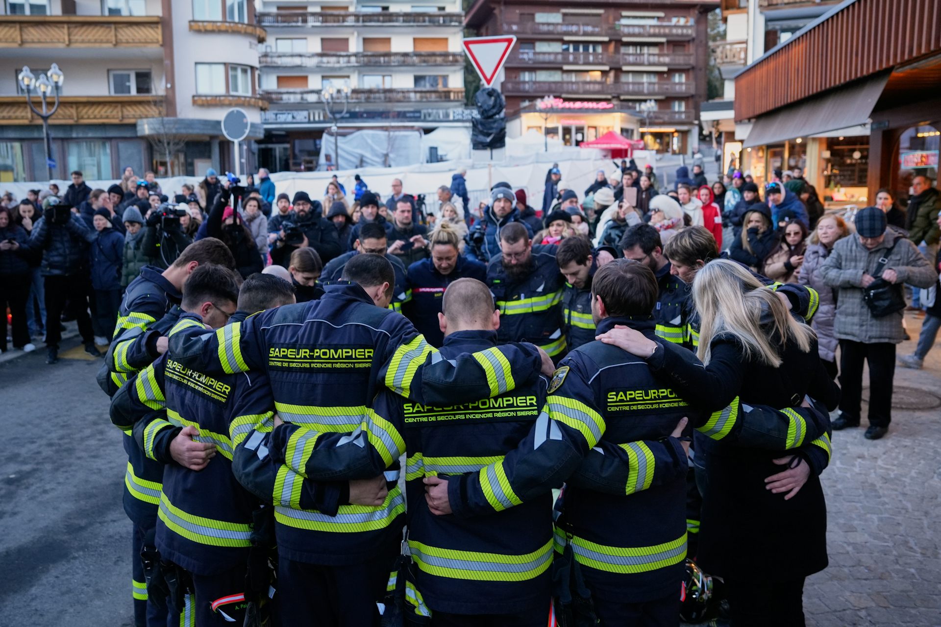 a group of firefighters in gear stand in a circle with their arms round each other. A crowd of other people in the background