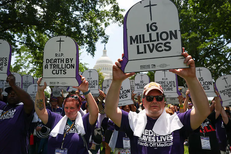 Protesters standing in front of a federal building, holding signs in the shape of graves reading '16 MILLION LIVES' and 'R.I.P. DEATH BY A THOUSAND CUTS,' wearing shirts that read 'MEDICAID SAVES LIVES'